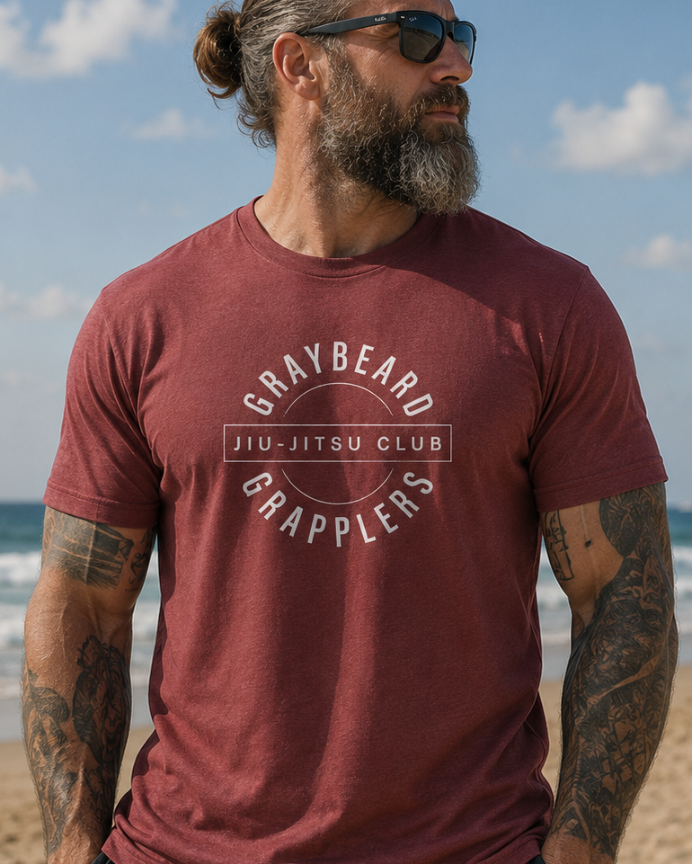 Man wearing a maroon t-shirt with 'Graybeard Jiu-Jitsu Club' text on a beach.